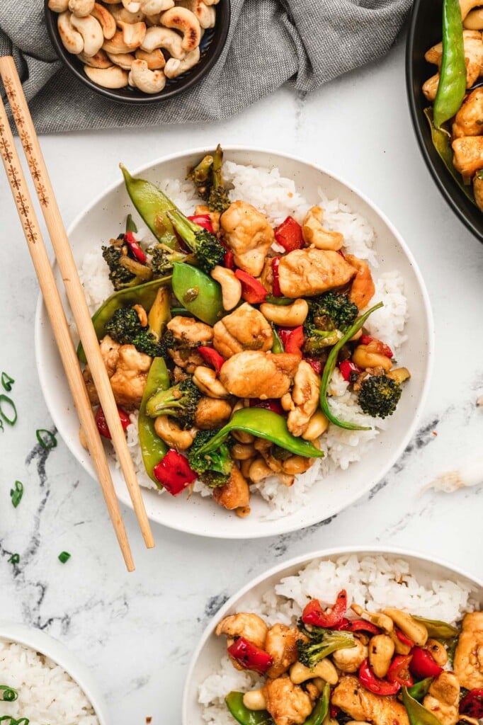 An overhead shot of a white plate with cashew chicken on a bed of white rice with chopsticks, another plate and a pan of cashew chicken on the side for one pot meals