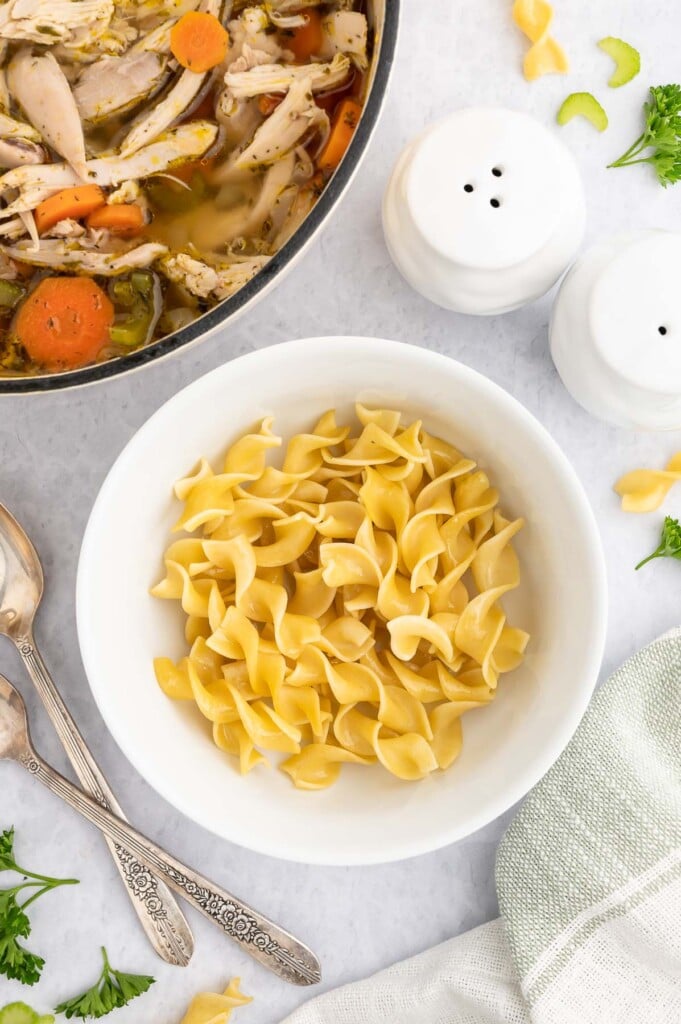 A white bowl of egg noodles with a dutch oven of rotisserie chicken soup on the side