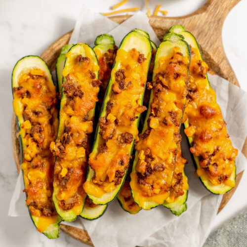 Stuffed Zucchini Boats on a pieces of parchment paper on a wooden cutting board with shredded cheese on the side