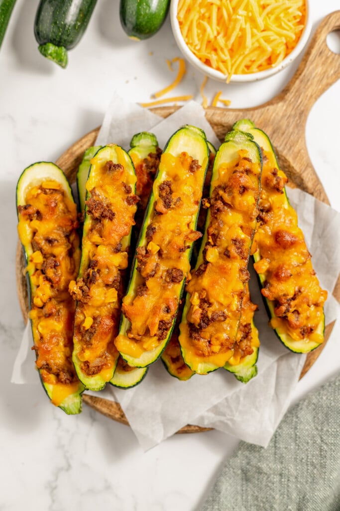 Stuffed Zucchini Boats on a pieces of parchment paper on a wooden cutting board with shredded cheese on the side