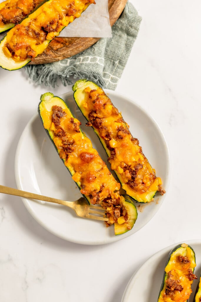Two stuffed zucchini boats on a white plate. One has a bite taken out with a gold fork holding it on the same plate with more stuffed zucchinis on the sides on a plate and platter.