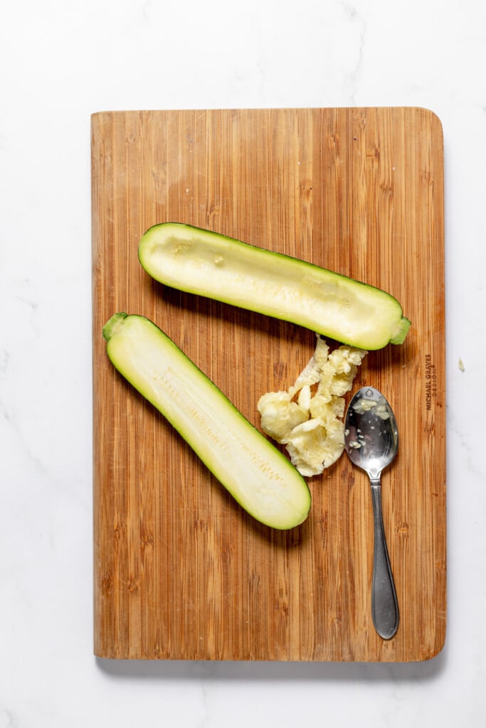A zucchini split in half on a wooden cutting board with the seedy center scooped out and set on the side with a spoon.