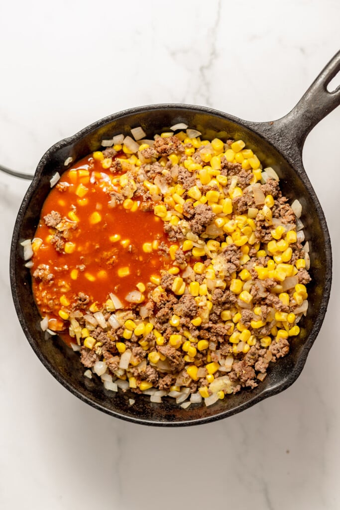 Red enchilada sauce being added to a ground beef filling in a cast iron pan for zucchini boats.