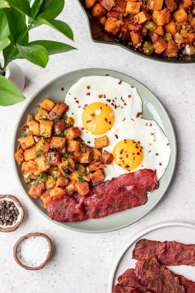 An overhead shot of Sweet Potato Home Fries on a grey plate with bacon and sunny side up eggs with the skillet of home fries on the side