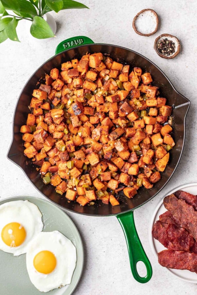 An overhead shot of sweet potato home fries in a skillet with sunny side eggs and bacon on plates on the side