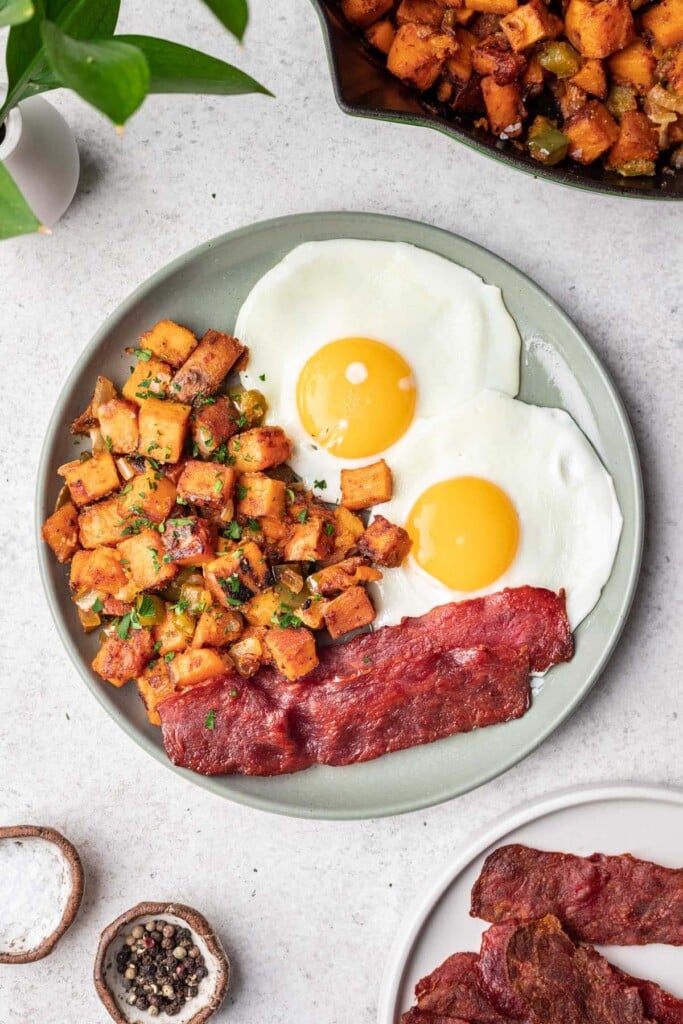 An overhead shot of Sweet Potato Home Fries on a grey plate with bacon and sunny side up eggs with the skillet of home fries on the side
