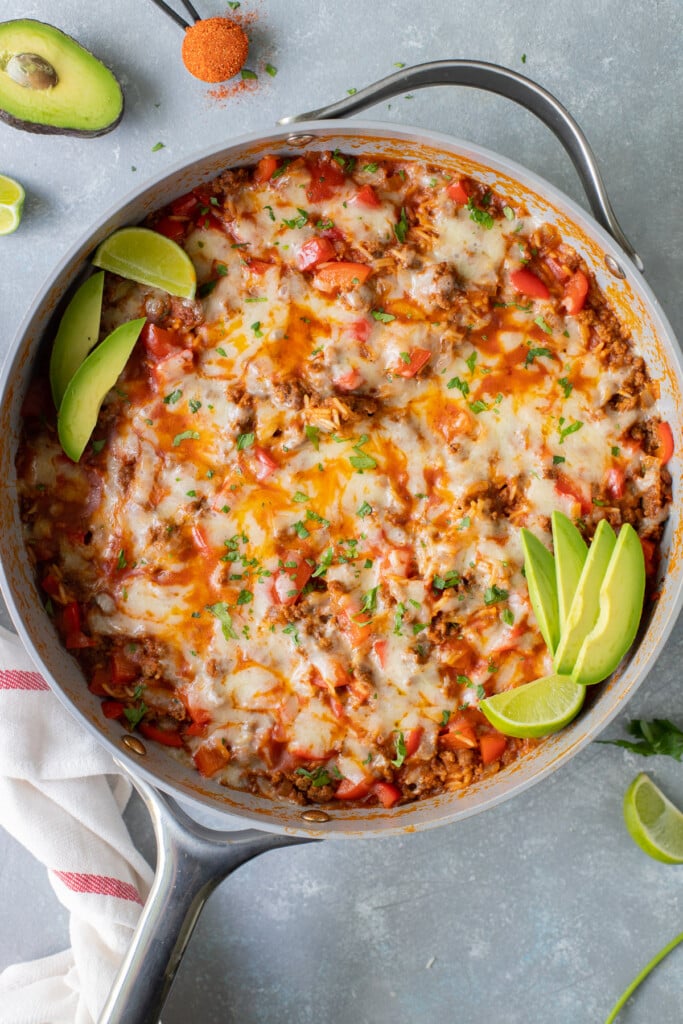 An overhead shot of a taco skillet in a large pan garnished with avocado slices and lime wedges on a grey background