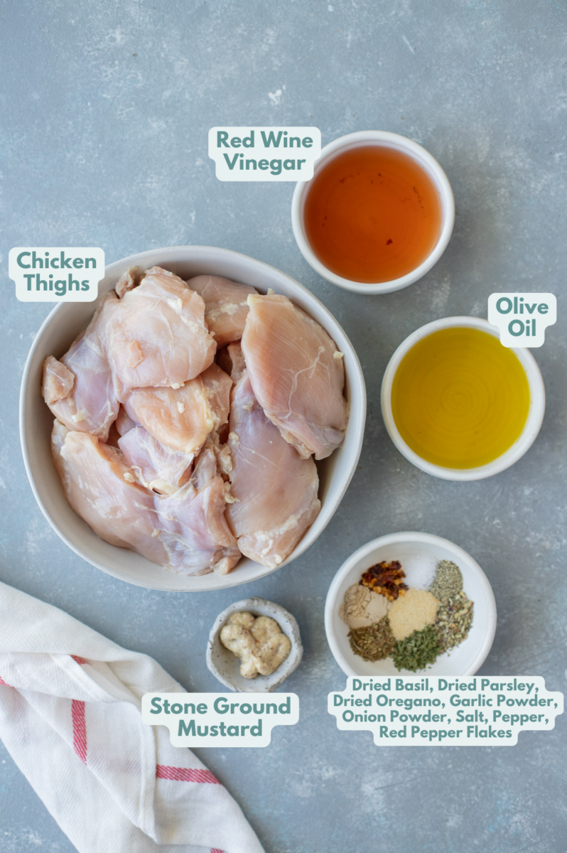 Ingredients for grilled chicken thighs in small bowls: boneless chicken thighs, olive oil, red wine vinegar, stone ground mustard, dried basil, dried parsley, dried oregano, garlic powder, onion powder, salt, pepper, red pepper flakes