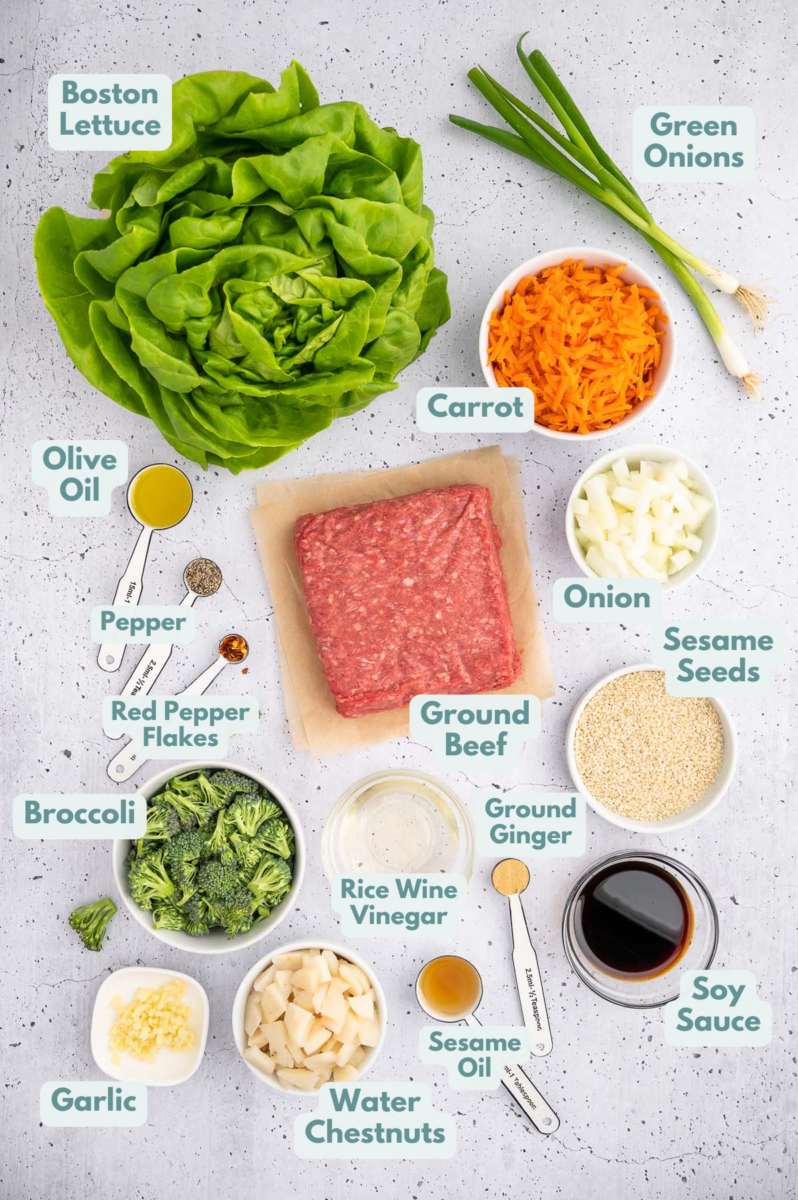 Ingredients for ground beef lettuce wraps in small bowls: olive oil, ground beef, onion, garlic, carrot, broccoli, water chestnuts, soy sauce, rice wine vinegar, sesame oil, pepper, ginger, red pepper flakes, boston lettuce, sesame seeds, green onions