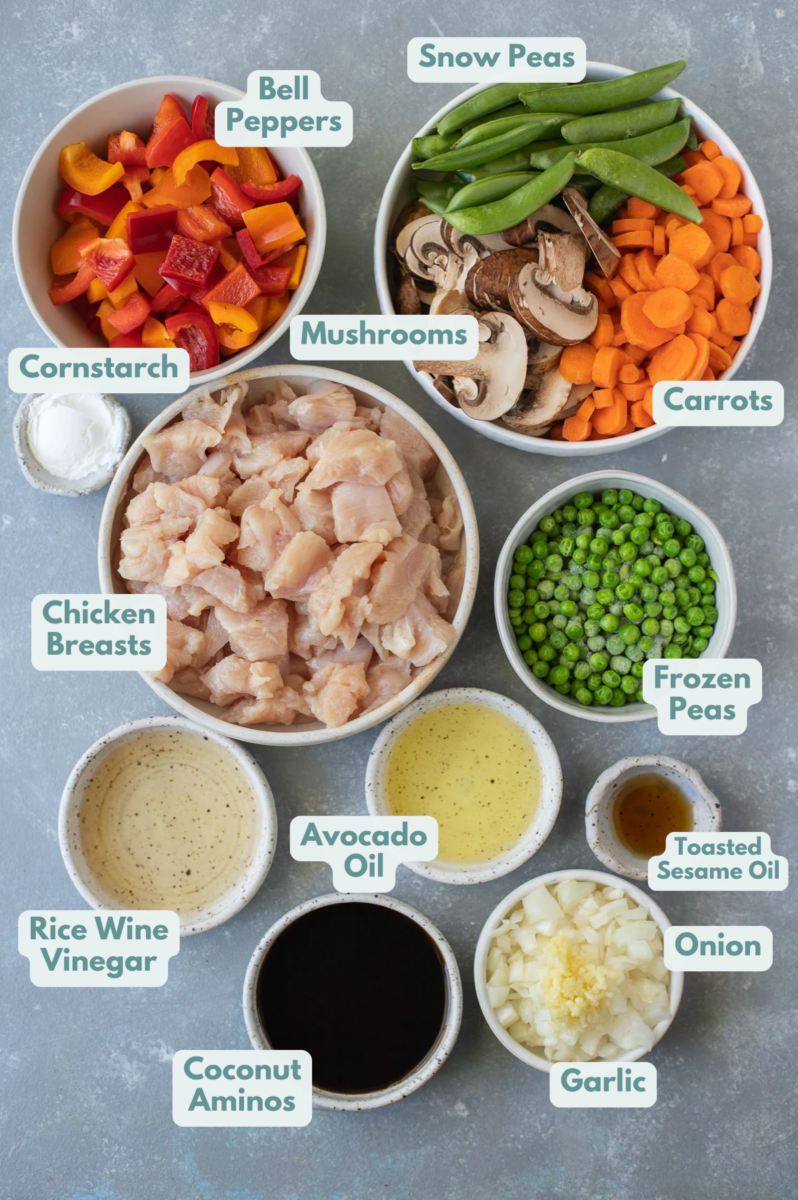 Ingredients for chicken stir fry in small bowls on a grey background: avocado oil, chicken breasts, rice wine vinegar, cornstarch, onion, garlic, bell peppers, carrots, mushrooms, snow peas, frozen green peas, coconut aminos, rice wine vinegar, toasted sesame oil, lime juice, honey, cooked white rice
