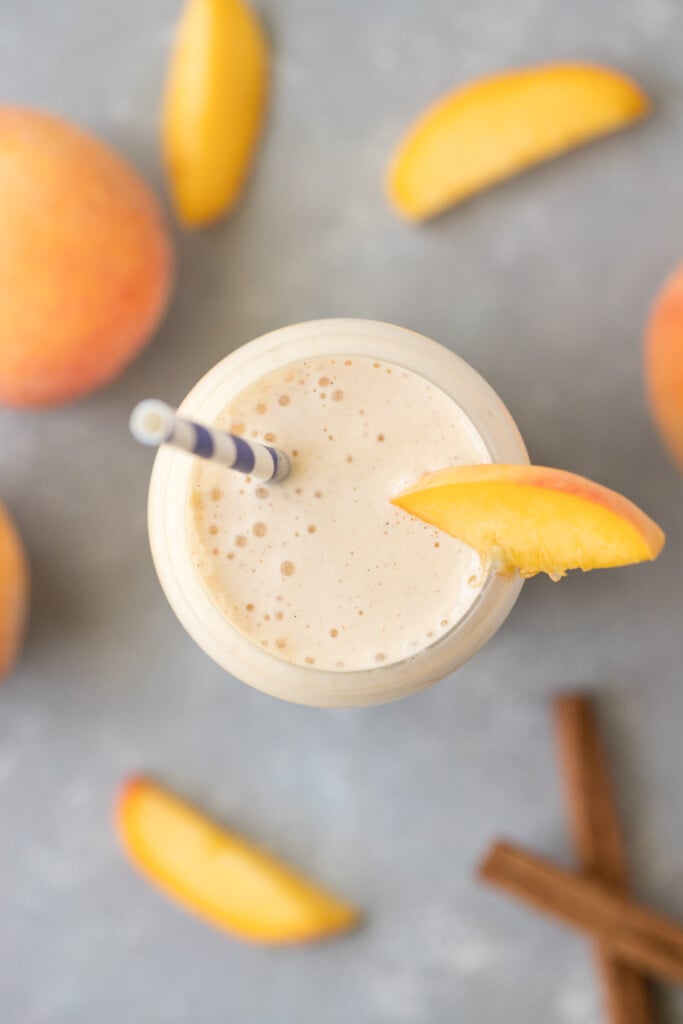 An overhead shot of peach smoothie in a clear glass with a blue and white striped straw in it garnished with a peach slice and more sliced peaches and cinnamon sticks on the side