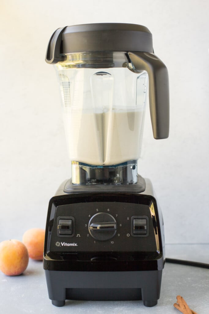 A vitamix blender with a peach smoothie in it with peaches in the background