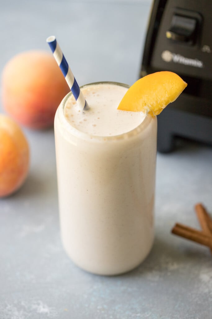 A peach smoothie in a clear glass with a blue and white striped straw in it garnished with a peach slice, a vitamix and more peaches in the background