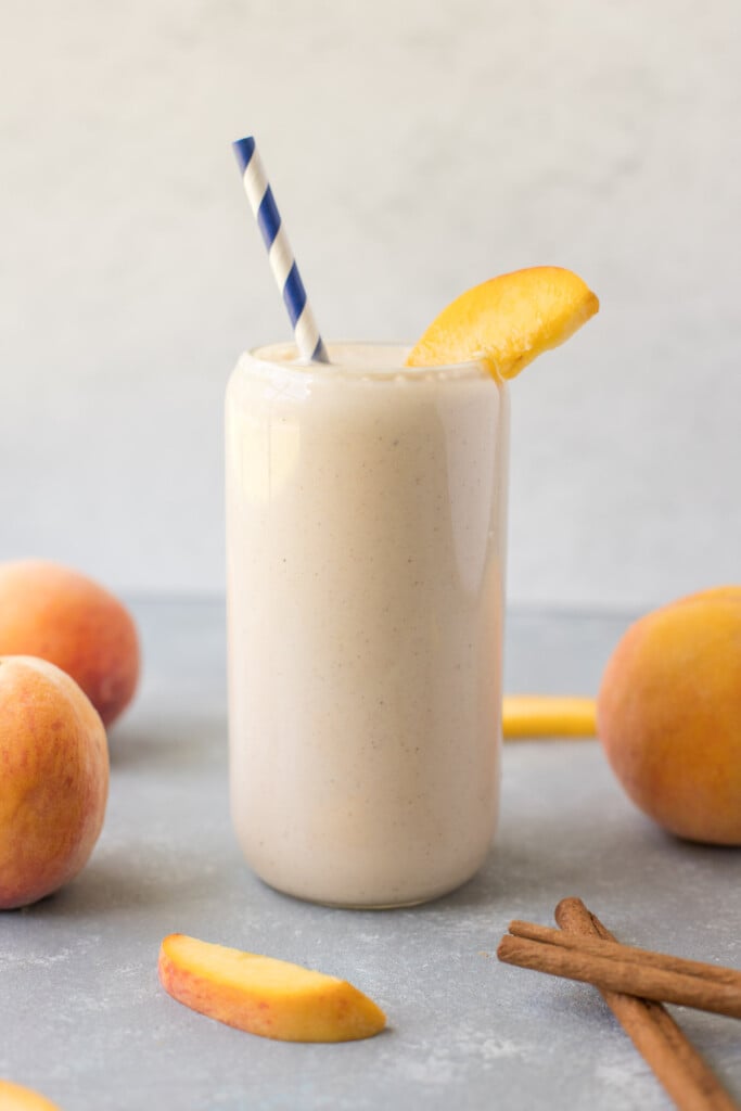 A peach smoothie in a clear glass with a blue and white striped straw in it garnished with a peach slice and more sliced peaches and cinnamon sticks on the side