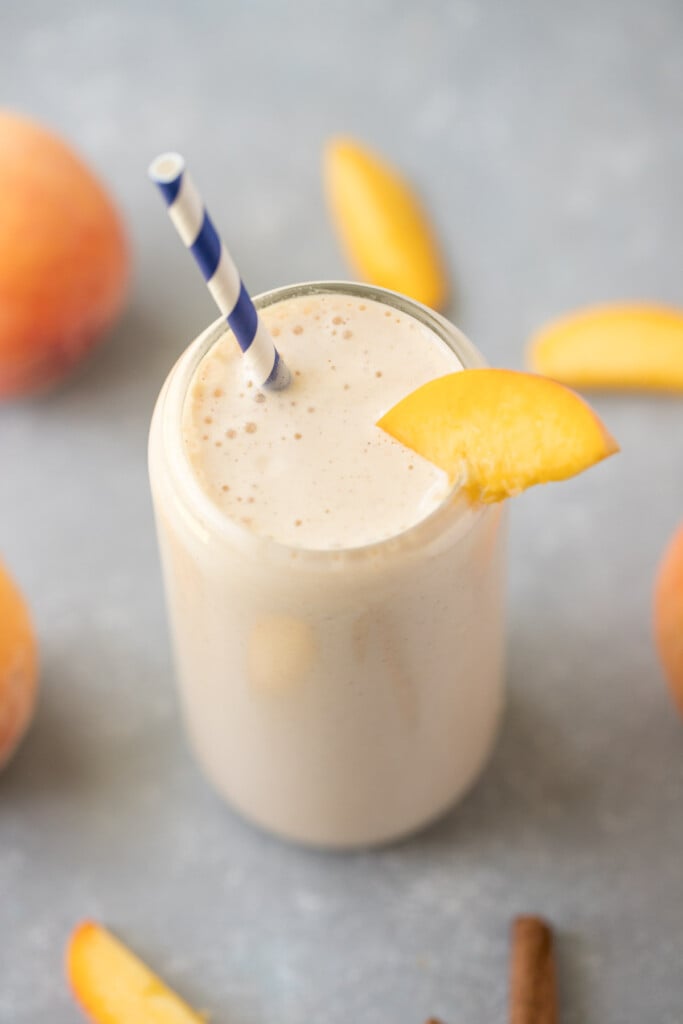 A peach smoothie in a clear glass with a blue and white striped straw in it garnished with a peach slice and more sliced peaches in the background