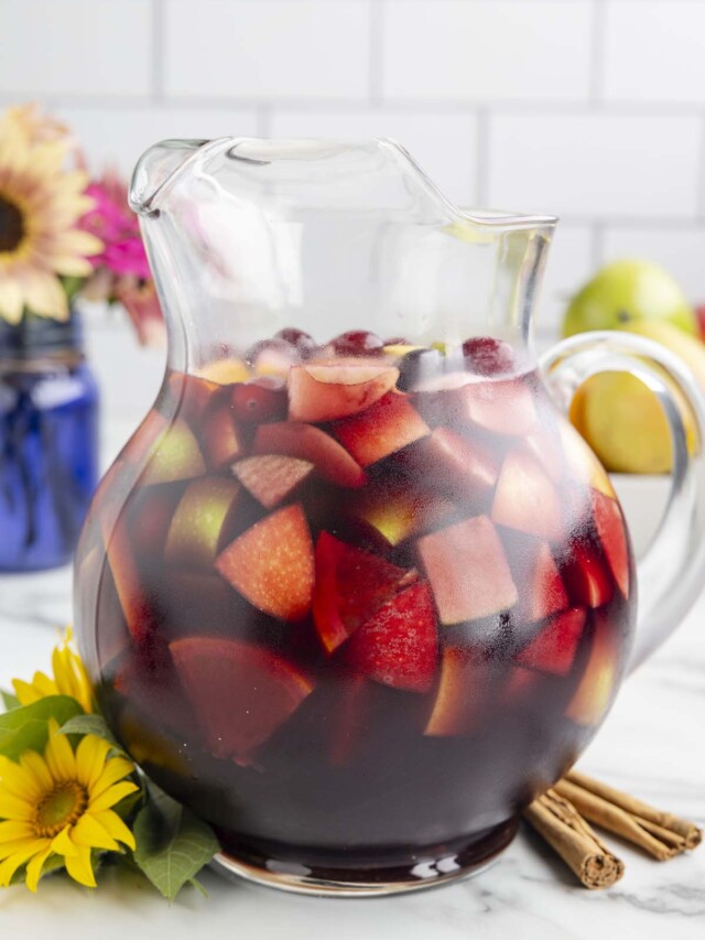 A clear glass pitcher filled with red wine sangria that has cranberries, apples and pears in it with flowers in the background