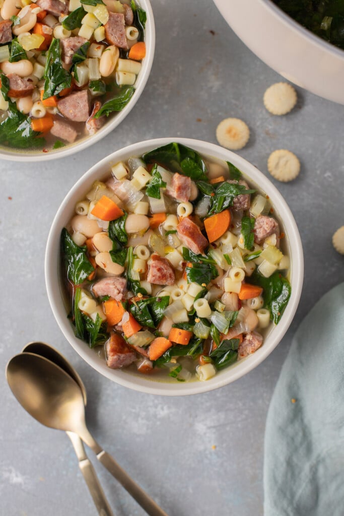 An overhead shot of a bowl of polish sausage soup with another bowl of soup and spoons on the side on a grey backgroud