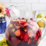 A clear glass pitcher filled with red wine sangria that has cranberries, apples and pears in it with flowers in the background