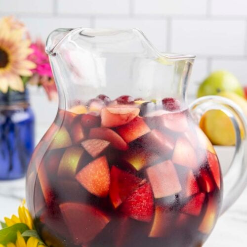 A clear glass pitcher filled with red wine sangria that has cranberries, apples and pears in it with flowers in the background