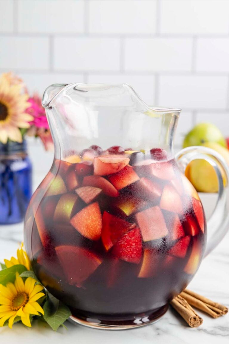 A clear glass pitcher filled with red wine sangria that has cranberries, apples and pears in it with flowers in the background