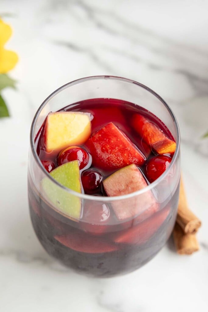 A clear glass with red sangria that contains fruit like cranberries, pear, apple, oranges on a marble counter