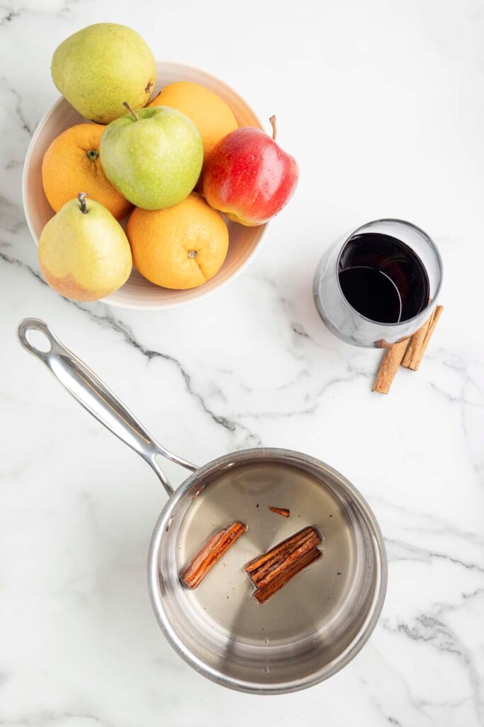 A saucepan of cinnamon simple syrup with red wine glass and a bowl of fruit (red and green apples, oranges, pear) on a marble counter