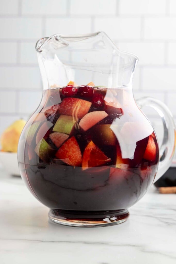 A clear glass pitcher filled with red wine sangria that has cranberries, apples and pears in it with a white background