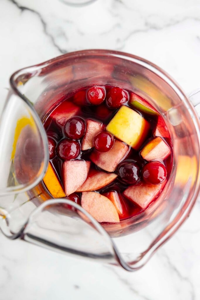 An overhead shot of red wine sangria recipe in a clear glass pitcher on a marble background