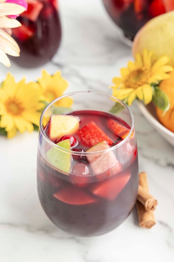 A clear glass with red sangria that contains fruit like cranberries, pear, apple, oranges on a marble counter with cinnamon sticks and flowers on the side