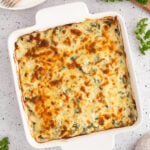 An overhead of a spinach artichoke chicken bake in a white casserole dish with a white plate with forks on top on the side on a white counter.