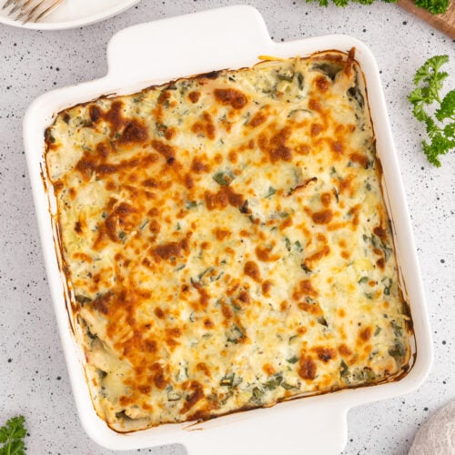 An overhead of a spinach artichoke chicken bake in a white casserole dish with a white plate with forks on top on the side on a white counter.