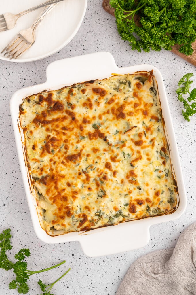 An overhead of a spinach artichoke chicken bake in a white casserole dish with a white plate with forks on top on the side on a white counter.