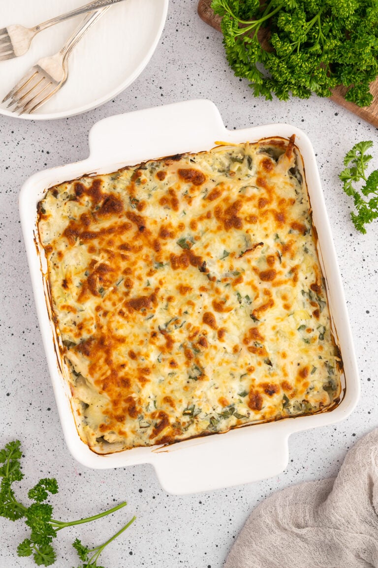 An overhead of a spinach artichoke chicken bake in a white casserole dish with a white plate with forks on top on the side on a white counter.