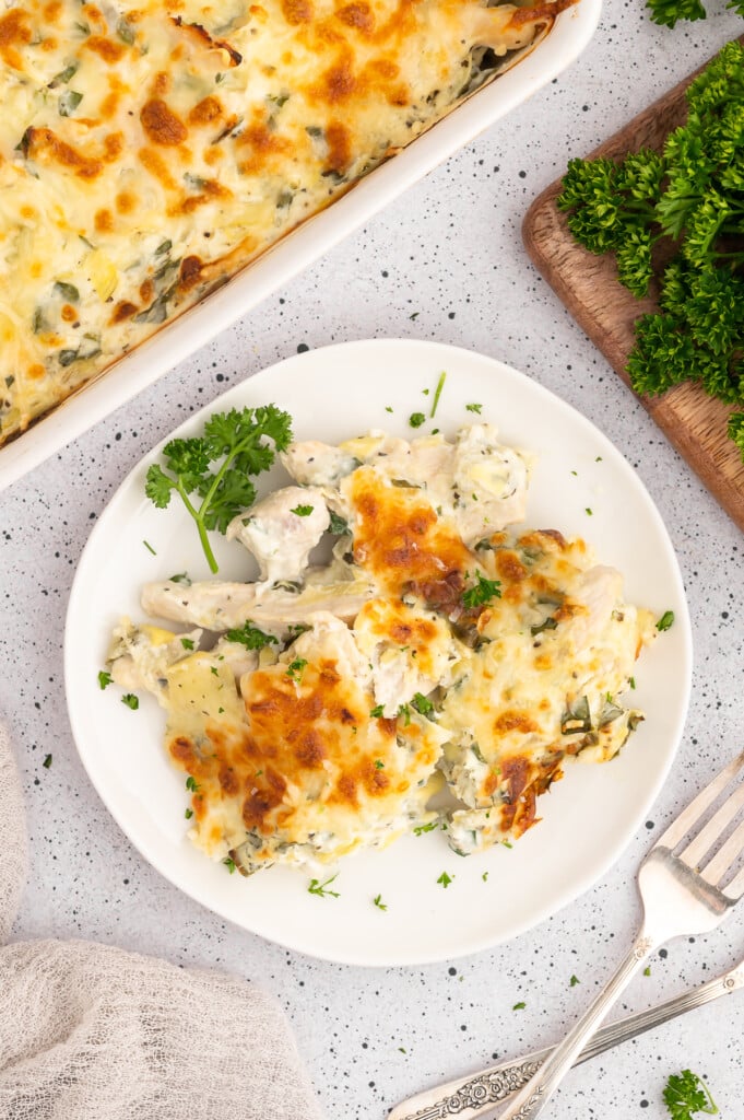A serving of spinach artichoke chicken on a white plate with the baking dish of the casserole on the side on a white counter.