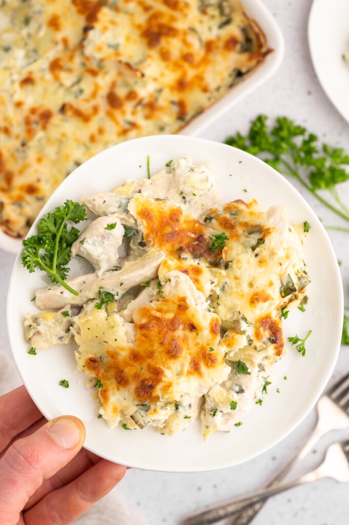 A hand holding up a plate with a serving of spinach artichoke chicken and the baking dish of the casserole in the background.