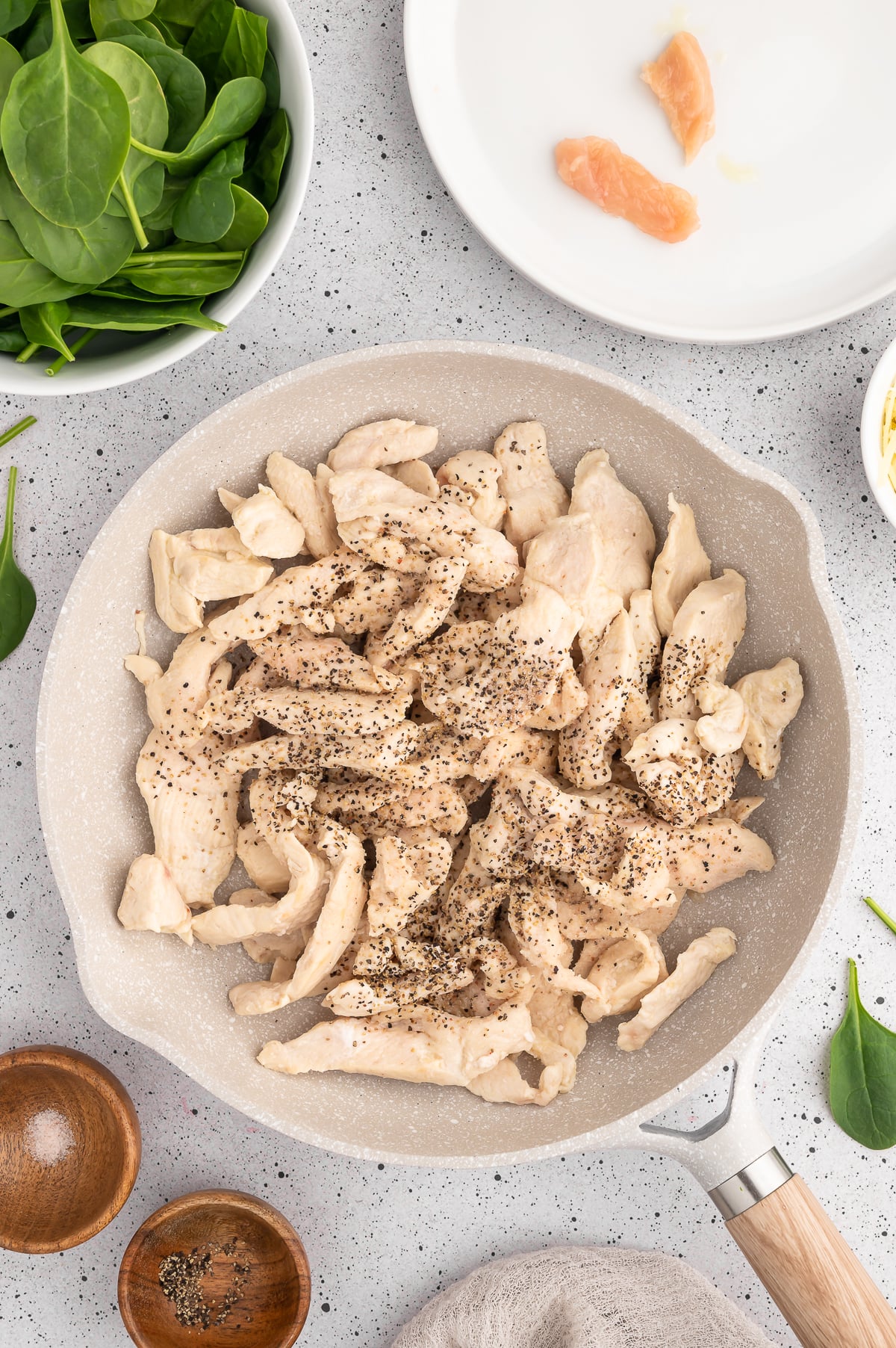 Chicken pieces in a skillet cooked and topped with salt and pepper with ingredients for spinach artichoke chicken on the side.