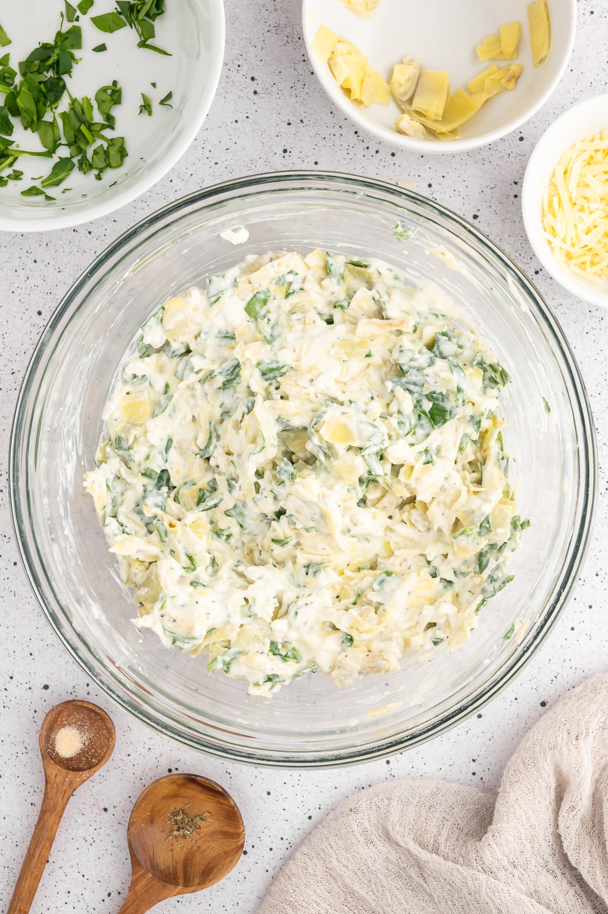 Cream cheese, spinach, artichoke mixture for spinach artichoke chicken bake in a clear mixing bowl on a grey counter.