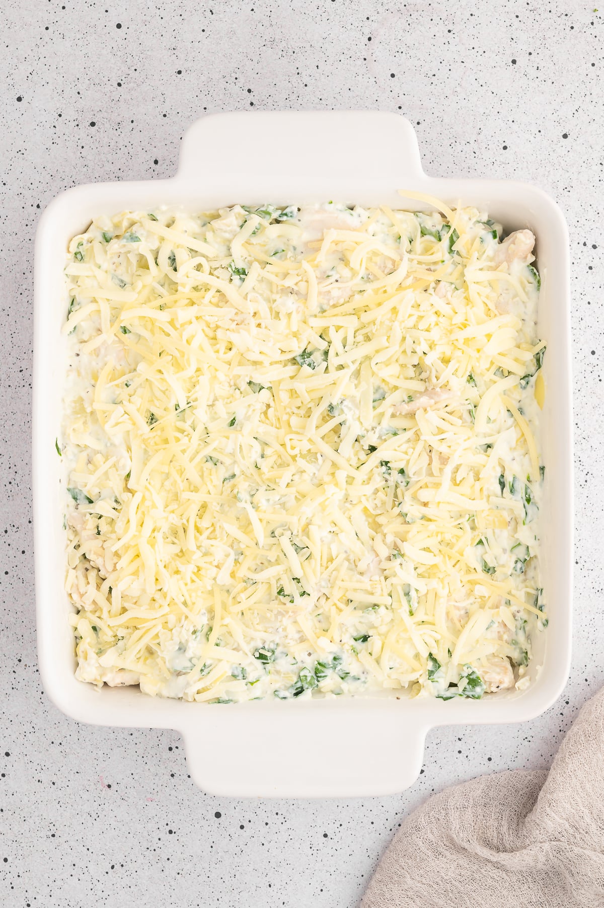 Uncooked spinach artichoke chicken bake in a white baking dish on a grey counter.