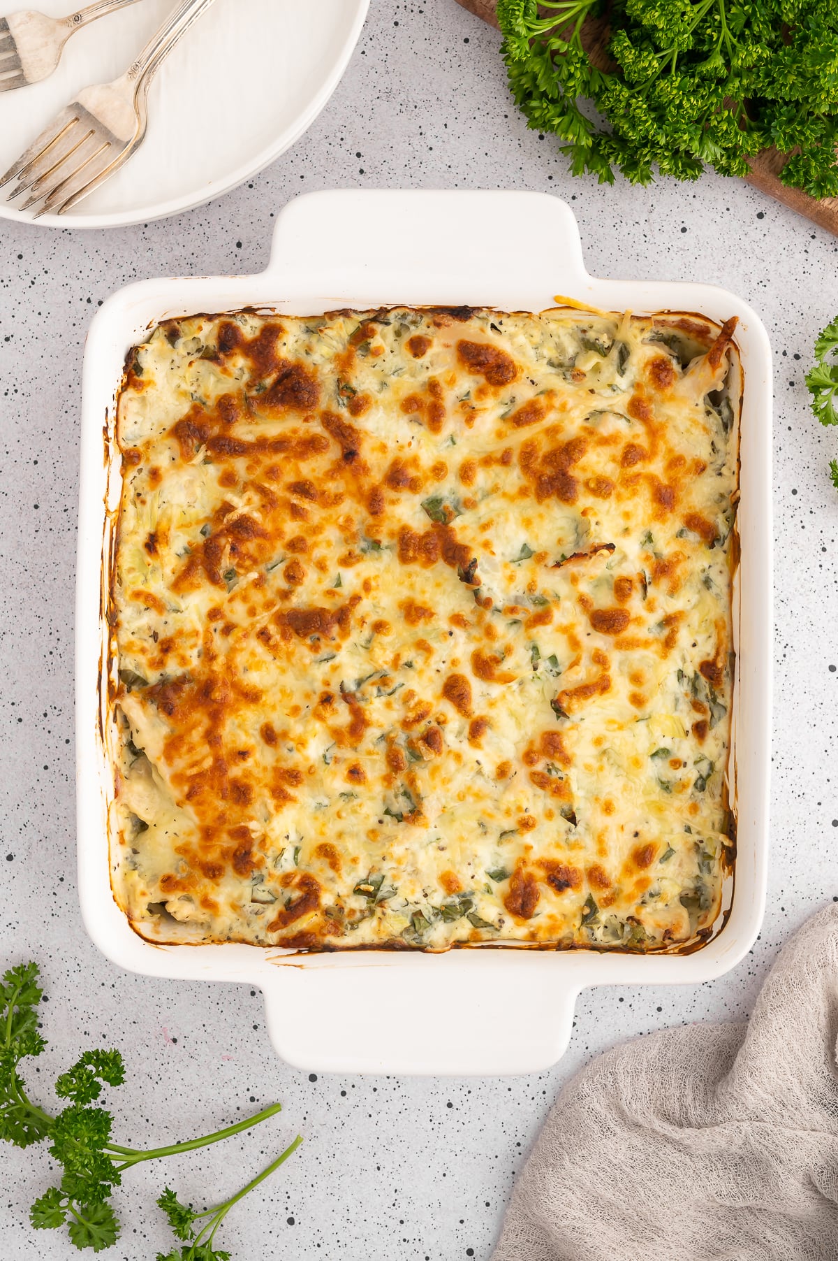 Spinach Artichoke chicken bake in a white baking dish with fresh herbs on the side on a grey counter.