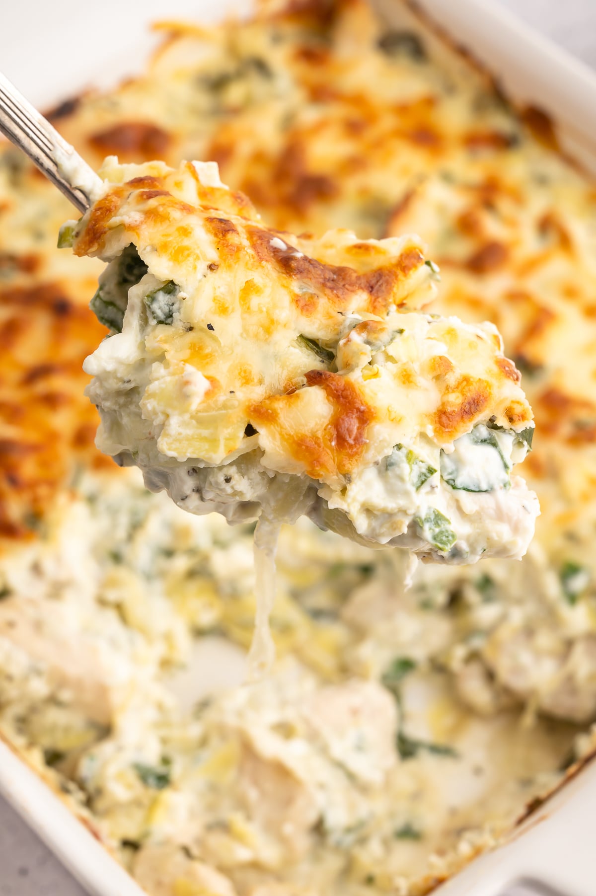 A large spoon holding up a scoop of spinach artichoke chicken with the baking dish in the background.