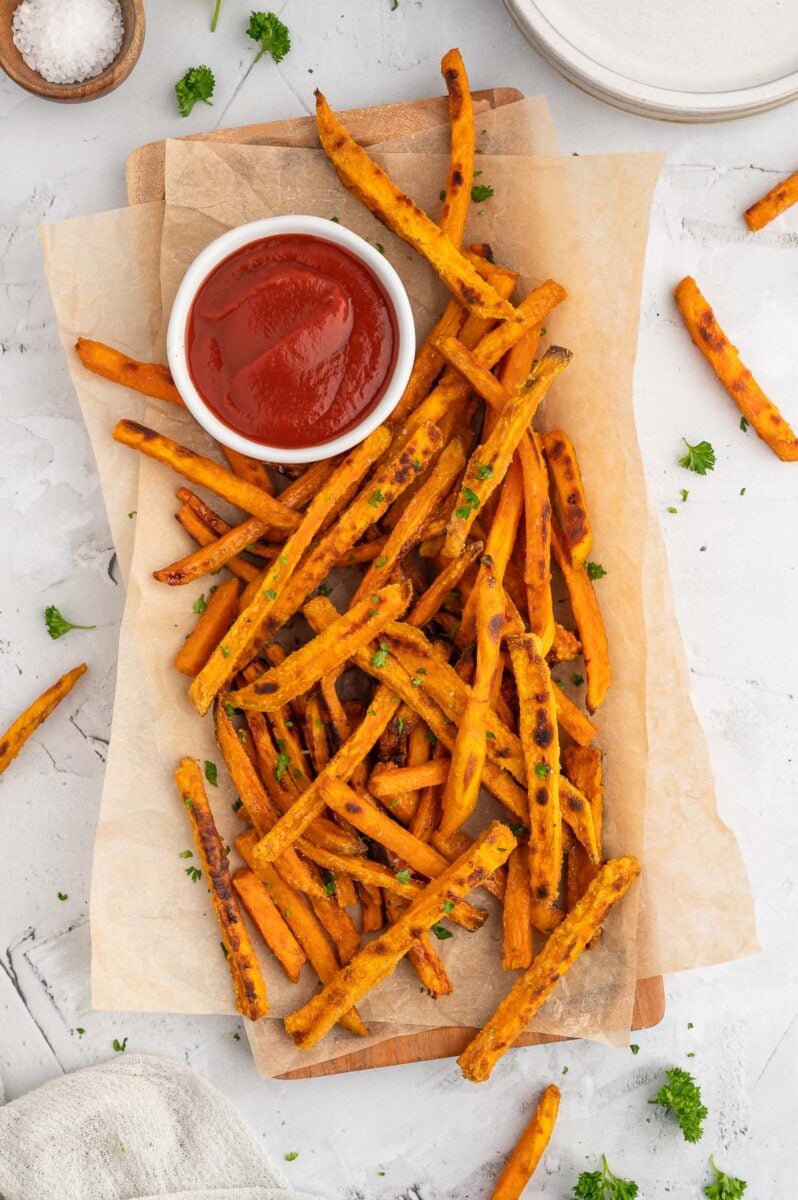 An overhead shot of baked sweet potato fries on a parchment lined platter with a white bowl of ketchup on the platter on a grey background