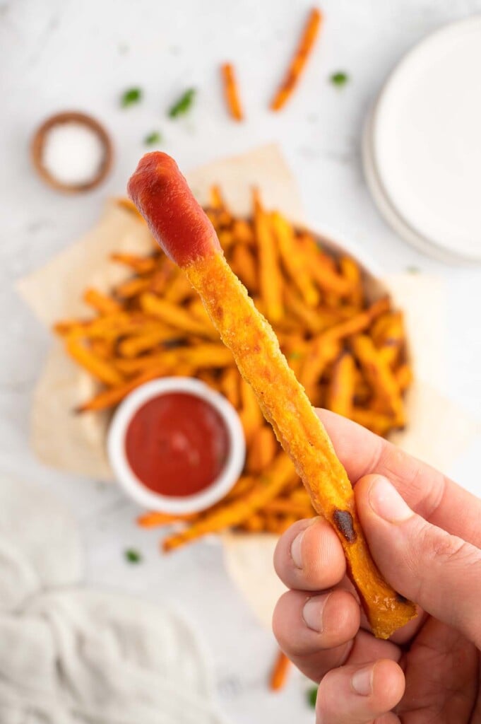 A hand holding up a single crispy sweet potato fry that has been dipped in ketchup with a platter of the oven baked sweet potato fries in the background