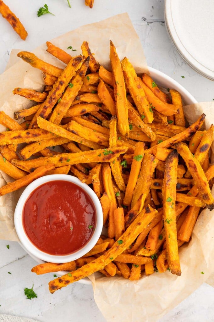 An overhead shot of crispy sweet potato fries in a round platter that is lined with parchment paper with a white bowl of ketchup on the platter