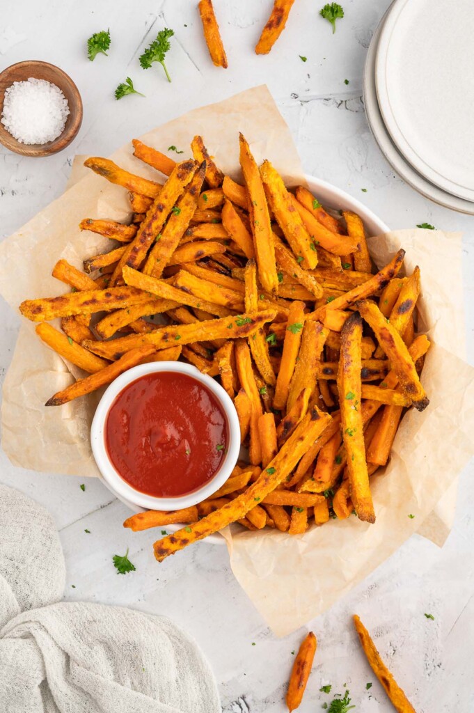 An overhead shot of oven baked sweet potato fries in a round platter that is lined with parchment paper with a white bowl of ketchup on the platter