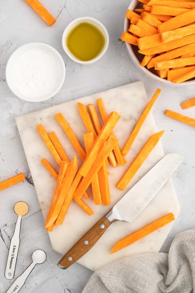 An overhead shot of uncooked sweet potato fries and a knife on a marble cutting board with a bowl of the  fries, olive oil, tapioca starch on the side