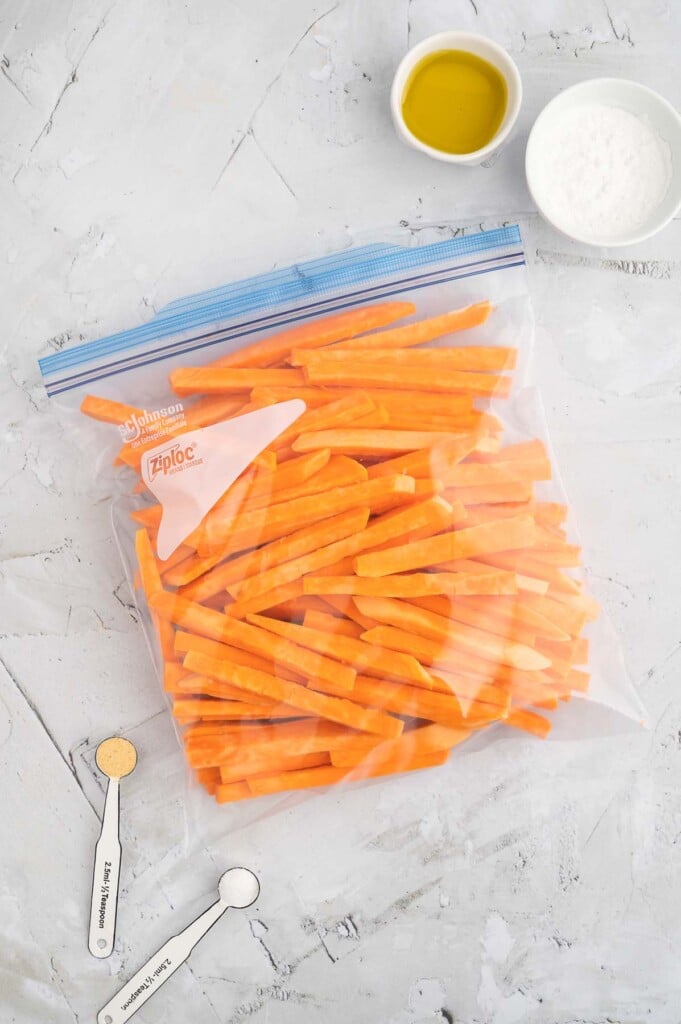 An overhead shot of fry sliced sweet potatoes in a ziploc bag with small bowls of olive oil, tapioca starch and spices on the side on a marble counter