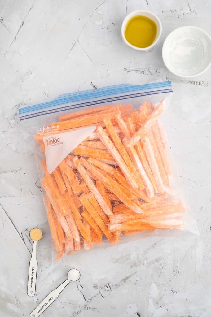 An overhead shot of fry sliced sweet potatoes tossed in tapioca starch in a ziploc bag with small bowls of olive oil and spices on the side on a marble counter