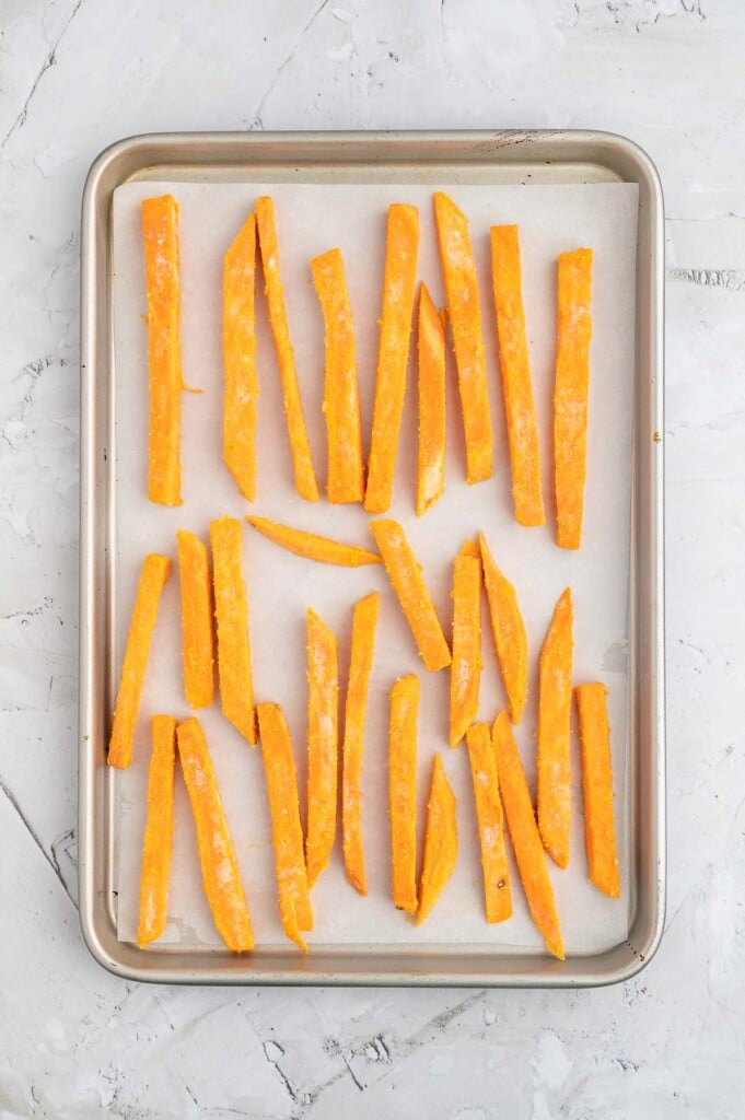 An overhead shot of a parchment lined baking sheet with unbaked sweet potato fries on a marble background