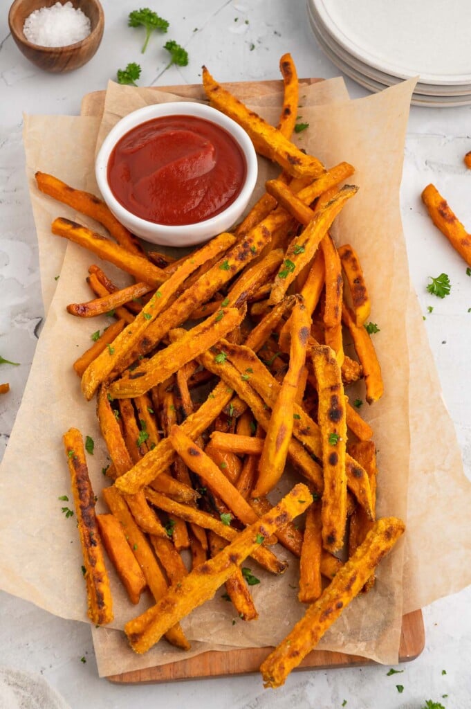 Sweet potato fries on a parchment lined platter with a white bowl of ketchup on the platter on a grey background