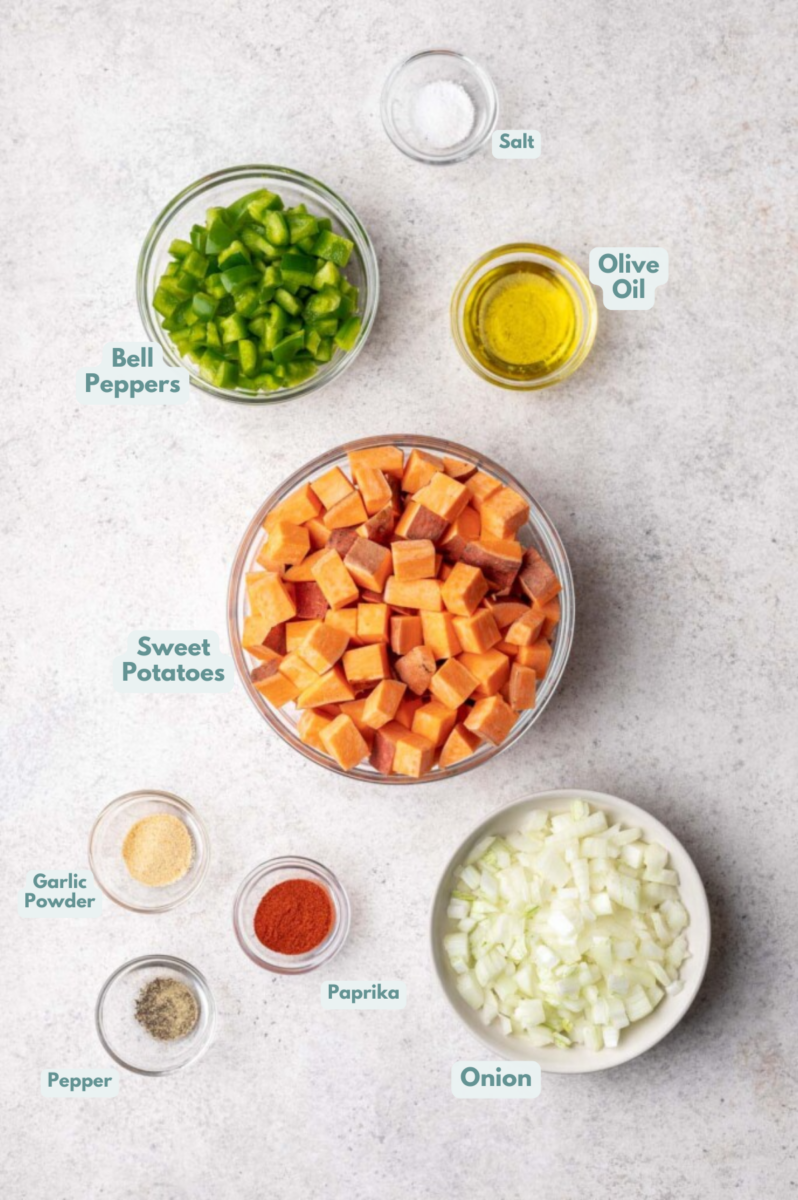 An overhead shot of ingredients for sweet potato home fries in small bowls: olive oil, onion, bell peppers, sweet potatoes, paprika, garlic powder, salt, pepper